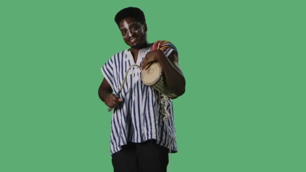 Portrait African American pria melihat kamera dan bermain african berbicara drum yuka. Laki-laki kulit hitam dengan pola etnis putih di wajah dan bergaris-garis kemeja nasional berpose di layar hijau di studio. — Stok Video