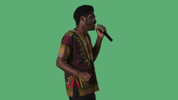 Portrait jeune homme afro-américain chantant une chanson au microphone. Homme noir avec un motif ethnique blanc sur le visage et en chemise lumineuse avec ornement national posant sur écran vert en studio. Vue latérale — Video