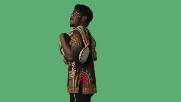Retrato joven afroamericano toca yuka de tambor africano parlante. Hombre negro con patrón étnico blanco en la cara y en camisa brillante con adorno nacional posando en pantalla verde en el estudio. Vista lateral — Vídeo de stock