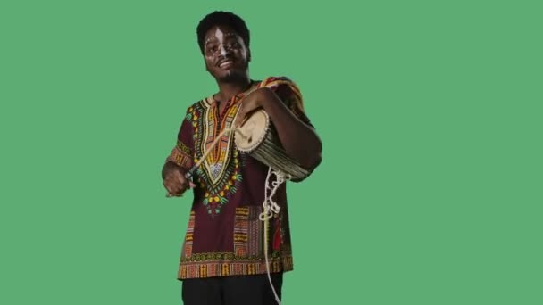 Porträt junger afroamerikanischer Mann spielt afrikanische sprechende Trommel Yuka. Schwarzer Mann mit ethnisch-weißem Muster im Gesicht und hellem Hemd mit nationalem Ornament posiert auf grünem Bildschirm im Studio. — Stockvideo