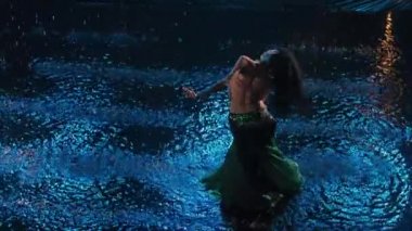 Karanlık stüdyoda mavi ışıkla su yüzeyinde göbek dansı yapan büyüleyici doğulu dansçının en iyi görüntüsü. Arap egzotik dansçı kalçalarını yağmur damlaları arasında ağır çekimde hareket ettirir..