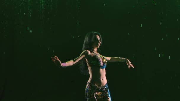 Dançarina oriental encantadora dança apaixonadamente uma dança exótica em revelar traje brilhante contra o fundo de quedas de chuva, fumaça e luz verde. Fecha. Silhueta. Movimento lento. — Vídeo de Stock