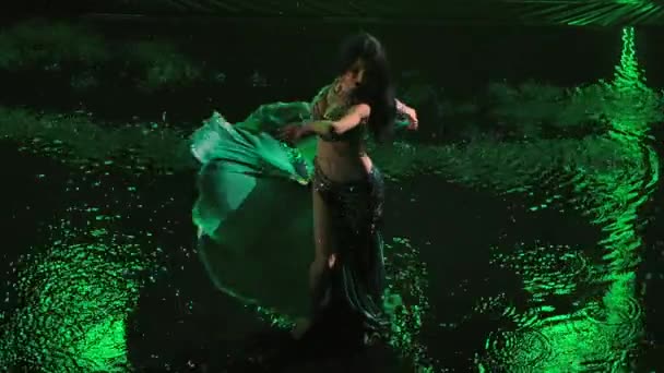 Ovanifrån av charmig orientalisk dansare som utför magdans stående på vattenytan i mörk studio med grönt ljus. Arabisk exotisk dansare flyttar höfterna i slow motion bland regndropparna. — Stockvideo