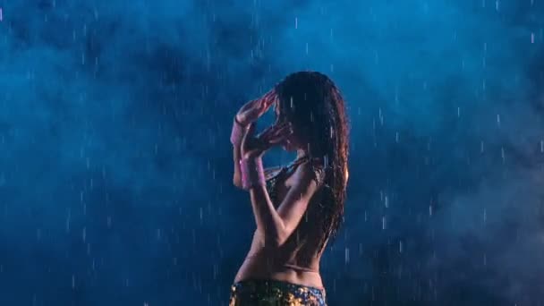 Silueta orientálního tance břicha a třesoucího se boky v dešti proti tmavému zakouřenému studiu s modrým světlem. Žena s atraktivním tělem tančí vášnivě a vlní mokré vlasy. Zavřít. Zpomalený pohyb