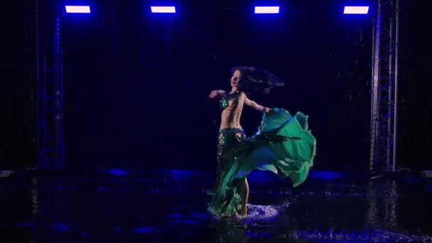 Young woman dances a belly dance, moves her hips, whirls and kicks water creating lot of splashes. An exotic oriental dancer performs among the raindrops in a dark studio with blue light. Slow motion. — Stock Video