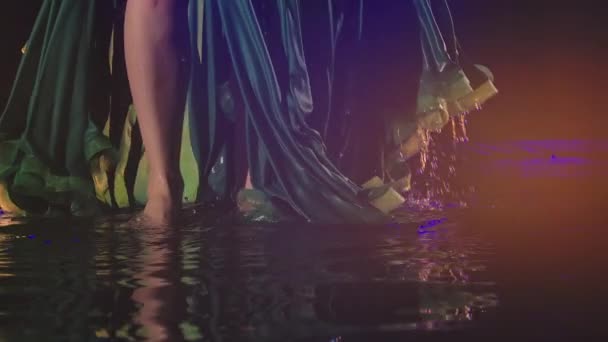 Close-up van een jonge oosterse dansers voeten raken het oppervlak van het water en het creëren van spatten. Vrouwelijke slanke poten in een lange groene rok op een donkere studioachtergrond. Langzame beweging. — Stockvideo