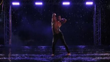 Modern balenin dans eden yakışıklı genç adamı, yağmur damlalarını bölüyor. Çıplak gövdeli erkek dansçı, karanlık stüdyoda suyun yüzeyinde su sıçramaları arasında plastikçe hareket eder. Yavaş çekim.