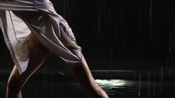 Mokra młoda kobieta w białej koszuli i beżowym body tańczy dramatyczny taniec nowoczesnego baletu w deszczu. Balet tancerz przenosi plastik, wykonując nowoczesną choreografię na wodzie powierzchniowej w ciemnym studio. Zamknij się. — Wideo stockowe