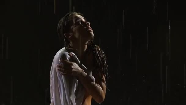 Beyaz gömlekli ve bej elbiseli ıslak genç kadın yağmurda dramatik modern bale dansı yapıyor. Bale, karanlık stüdyoda yüzeydeki su üzerinde modern koreografi yaparak plastiği hareket ettirir. Kapat. — Stok video