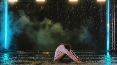 Genç bir kadın yağmurda suyun yüzeyinde otururken modern bale koreografisi yapıyor. Esnek bir kadın vücudu güzel bir ışıkla karanlık bir stüdyoda kıvranıyor. Yavaş çekim.