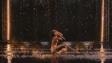 Genç kadın, akrobatik elementlerle koreografi modern bale yapıyor. Yağmurda su yüzeyinde vücut büküyor. Islak mayo giymiş seksi kadın silueti karanlık stüdyoda şehvetli bir şekilde hareket ediyor. Yavaş çekim.
