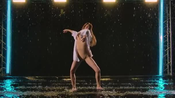 Hedendaagse sensuele ballet choreografie uitgevoerd door slanke flexibele ballerina temidden van de regendruppels. Natte vrouw in beige badpak dansend romantisch in donkere studio. Langzame beweging. — Stockvideo