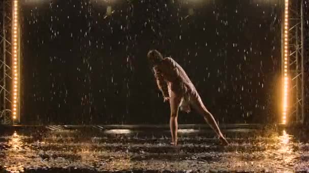 Coreografía sensual contemporánea de ballet realizada por una esbelta bailarina flexible en medio de las gotas de lluvia. Mujer mojada en traje de baño beige bailando románticamente en un estudio oscuro. Silueta. Movimiento lento. — Vídeos de Stock
