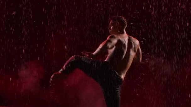 Sylwetka mężczyzny z nagim tułowiem wykonuje choreografię współczesnego baletu w deszczu na czarnym tle z czerwonym światłem. Mokry tancerz podczas tańca tworzy wiele plusków. Zwolnij trochę. Zamknij się.. — Wideo stockowe