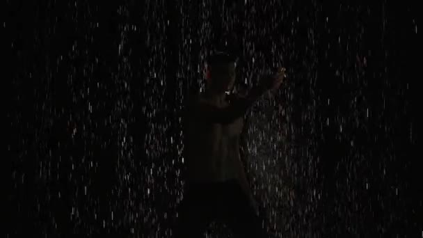 Silhouette of wet male dancer performing modern ballet dance in rain and splashing water. Male sensual modern choreography on black background in center of light beam. Slow motion. Close up. — Stock Video