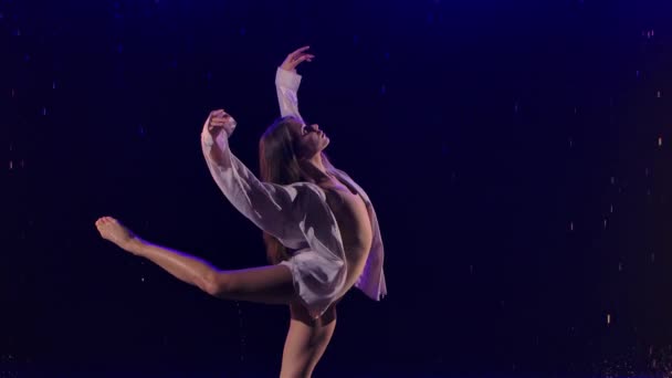 Mulher de camisa branca está dançando danças dramáticas de balé moderno na chuva. Dançarina de balé move plástico, realizando coreografia moderna em estúdio escuro com luz azul. Movimento lento. Fechar. — Vídeo de Stock