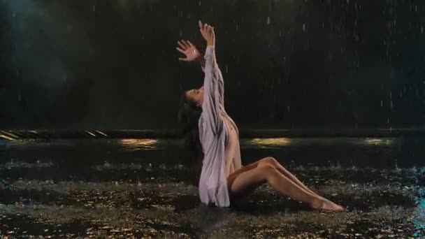 Uma jovem realiza coreografia moderna de balé enquanto se senta na superfície da água na chuva. Um corpo feminino flexível wriggles em um estúdio escuro com luz bonita. Movimento lento. Fechar. — Vídeo de Stock