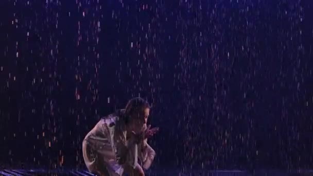 A bailarina de dança contemporânea dança sensualmente na chuva na superfície de água, criando salpicos. A silhueta da mulher molhada na camisa branca move-se plasticamente no estúdio escuro com a luz azul. Movimento lento. Fechar — Vídeo de Stock