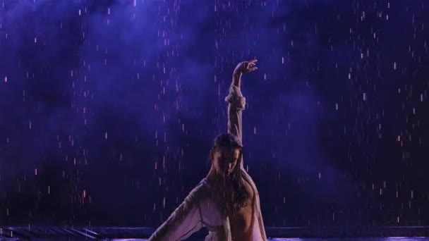 Bailarina de danza contemporánea baila sensualmente bajo la lluvia sobre la superficie del agua, creando salpicaduras. La silueta de la mujer mojada en la camisa blanca se mueven plásticamente en el estudio oscuro con la luz azul. En cámara lenta. De cerca. — Vídeos de Stock