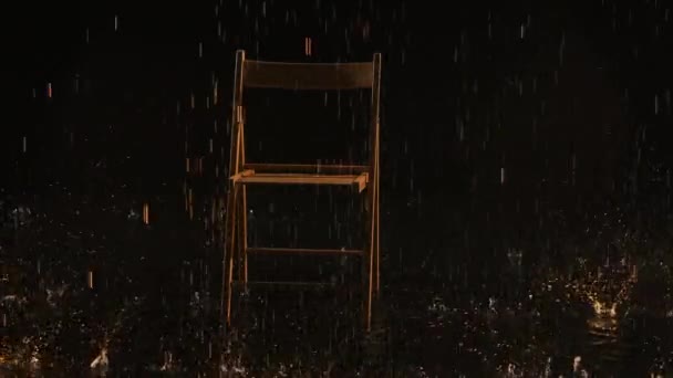 A wooden chair stands in the rain on the water surface in an empty dark studio. Raindrops illuminated by yellow light fall down and drum on the chair. Slow motion. Close up. — Vídeo de Stock