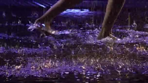 当代芭蕾舞演员在水面上的雨中灵敏地跳着舞,产生了水花.穿着白衬衫的湿湿的女人的轮廓在带有蓝光的黑暗工作室中可塑性地移动。慢动作靠近点 — 图库视频影像