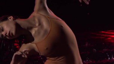 Yağmurda dans eden ince, esnek balerin silueti. Bej mayolu ıslak kadın kırmızı ışıkla aydınlatılmış yüzey suyunda yatarken şehvetli bir şekilde hareket ediyor. Kapatın. Yavaş çekim.