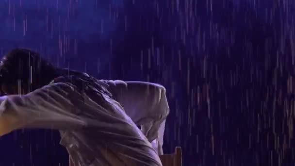 A bailarina de dança moderna contemporânea move-se sensualmente na chuva, sentada na cadeira. A silhueta de uma mulher molhada em uma camisa branca se move plasticamente em um estúdio escuro com luz azul. Movimento lento. Fechar. — Vídeo de Stock