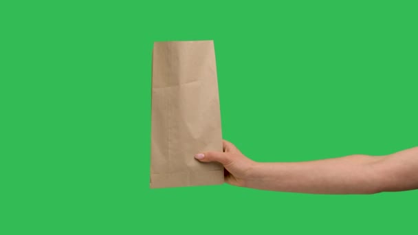 Female hand holds a paper shopping bag on the background of a green screen chroma key. Close up of paper packaging. Food and beverage courier delivery service concept. Advertising. Slow motion. — Stock video