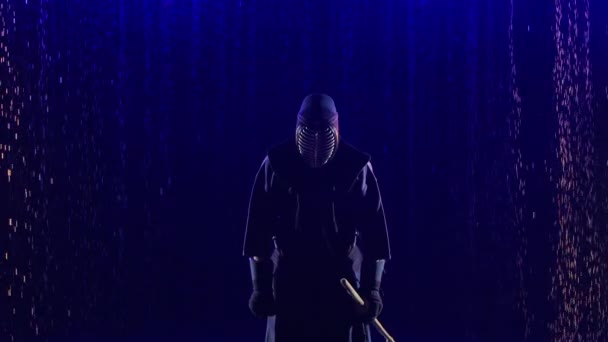 Japanska Kendo fighter sänka sin shinai mot kameran. Idrottare i kimono, kampsport och med bambusvärd poserar mot mörk studio bakgrund med blått ljus och regn. Närbild. Långsamma rörelser. — Stockvideo