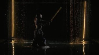 Japon kendo dövüşçüsü Shinai 'yi bıçakladı. Geleneksel savaş zırhlı ve elinde bambu kılıcıyla siyah stüdyo arka planı ve yağmura karşı duran bir adam. Yavaş çekim.