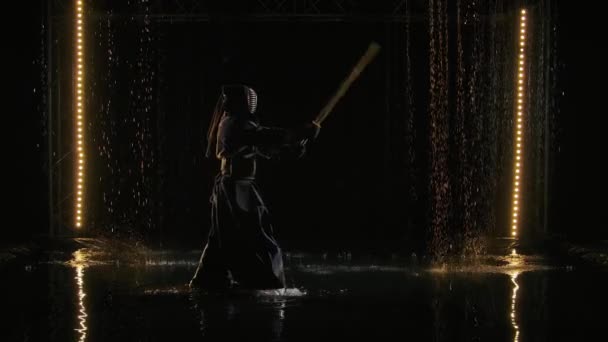 Japanese kendo fighter in fighting stance stabs with shinai. Man in traditional martial armor and with bamboo sword in hands poses against black studio background and rain. Slow motion. — Stock Video