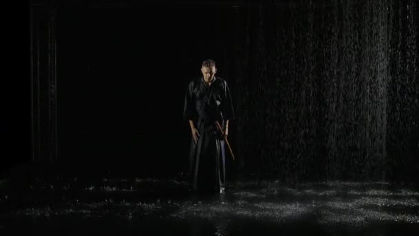 Luchador kendo japonés en posición de lucha sosteniendo su shinai. Hombre en kimono tradicional con espada de bambú marcial en sus manos posa sobre fondo de estudio negro bajo la lluvia. Movimiento lento. — Vídeos de Stock