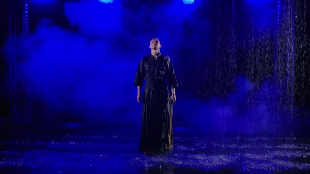 Luchador de kendo japonés practicando habilidad marcial bajo la lluvia sobre fondo de estudio ahumado oscuro con luz azul. El hombre está practicando golpes de combate balanceando su shinai. De cerca. Movimiento lento. — Vídeos de Stock