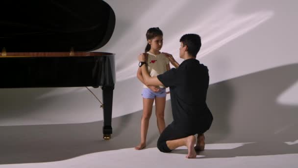 Persönliche Ballettstunde für kleines Mädchen. Professioneller Lehrer bringt der Ballerina Tanzelemente bei. Choreograph korrigiert Position der Hände und des Kopfes des Kindes auf weißem Studiohintergrund in der Nähe des Flügels — Stockvideo