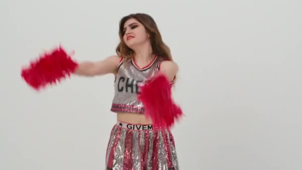 Young cheerleader with red pompoms in uniform is dancing on white background in studio. Beautiful cheerleader performs cheering dance. Pompons for cheerleading in female hands. Close up. Slow motion. — Stock Video