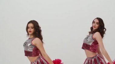 Amigo takım performansı. İki gülümseyen genç kadın senkronize bir şekilde dans ediyor ve kırmızı ponpon sallıyorlar. Parlak kostümlü ponpon kızların takım çalışması stüdyoda beyaz arka planda. Kapatın. Yavaş çekim.