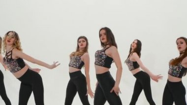 Gümüş bluzlu ve dar taytlı bir grup atletik ponpon kız beyaz stüdyo arka planında dans ediyorlar. Öğrenciler yavaş çekimde senkronize olarak dans edip ponpon sallıyorlar. Kapat..