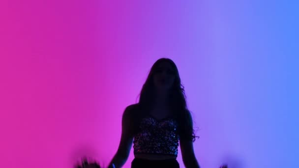 Silhouette einer schlanken Cheerleaderin, die energisch mit Pompons in den Händen vor dem Hintergrund rosa blauer Lichter im Studio tanzt. Jubeltanz bei Sportwettkämpfen. Aus nächster Nähe. Zeitlupe. — Stockvideo