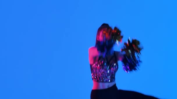 Energetic cheering dance performed by cute cheerleader with pompoms in hands against blue background, illuminated with pink light. Cheerleading, cheering sports team. Close up. Slow motion. — Stock Video