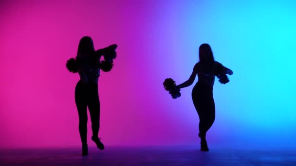 Silueta dvou štíhlých roztleskávaček, energicky tančících s bambulkami v rukou na pozadí růžově modrých světel ve studiu. Cheering dance ve sportovních soutěžích. Zpomalený pohyb. — Stock video