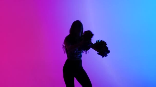 Silueta de esbelta animadora, bailando enérgicamente con pompones en las manos sobre fondo de luces azules rosadas en el estudio. Animando a bailar en competiciones deportivas. Movimiento lento. — Vídeos de Stock