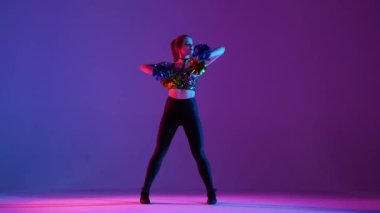 Atletik ince vücutlu genç bir kadın amigoluk yapıyor mavi mor bir stüdyo arka planında ponponlarıyla dans ediyor. Amigo kız ellerini sallıyor ve kalçalarını oynatıyor. Yavaş çekim.