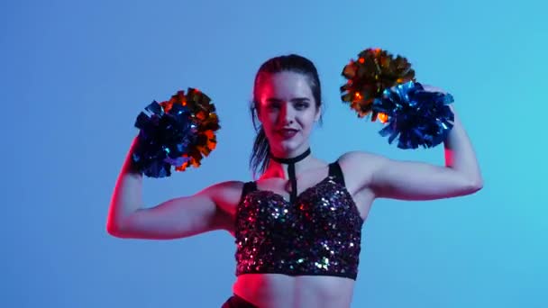 Retrato de una joven atractiva animadora bailando animando danza con pompones en las manos. Muchacha atractiva con cuerpo deportivo iluminado por luces azules rosadas. De cerca. Movimiento lento. — Vídeos de Stock