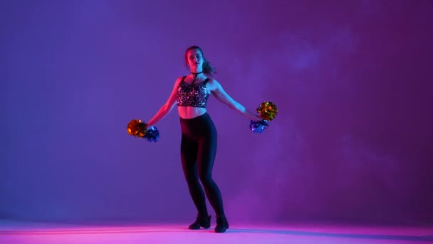 Young woman with an athletic slim body is practicing cheerleading, dancing with pompoms on a purple studio background with smoke. Cheerleader waves hands and sensually moves her hips. Slow motion. — Stock Video