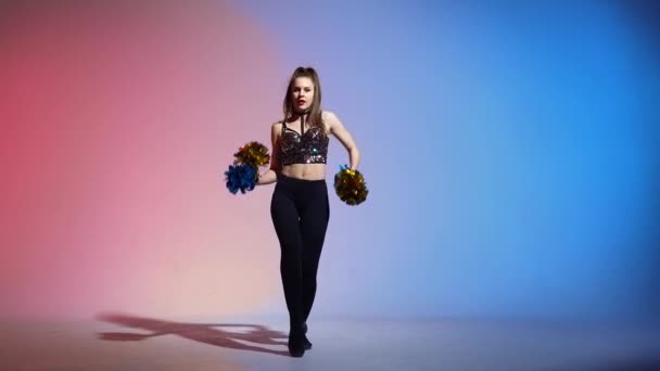 Energetic cheering dance performed by cute cheerleader with pompoms in hands against blue pink studio background. Adorable female moves hands and hips, whirls and waves hair. Slow motion. — Stock Video