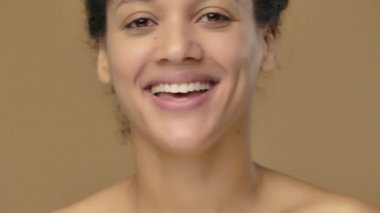 Afro-Amerikalı genç bir kadının güzel portresi gülümsüyor ve cilveli bir şekilde göz kırpıyor. Kahverengi stüdyo arka planında poz veren siyah bir kadın model. Kapatın. Yavaş çekim hazır, 4K 59.94fps 'de..