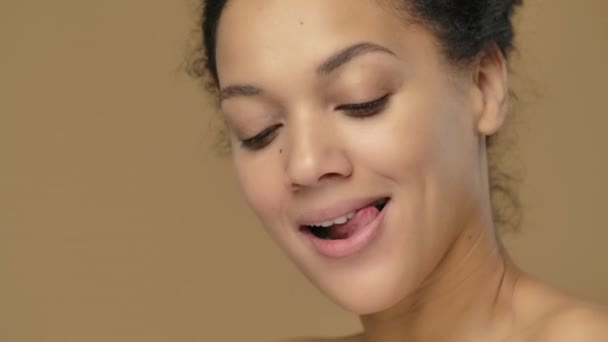 Красота портрет молодой афроамериканской женщины со здоровой кожей, смотрящей в камеру и кокетливо улыбающейся. Черная модель позирует на фоне коричневой студии. Закрывай. Медленное движение готово, 4K на 59,94fps — стоковое видео