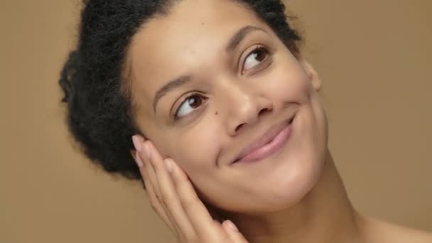 Retrato de belleza de una joven afroamericana que sueña con algo agradable y sonriente dulcemente. Modelo femenino negro posando sobre fondo de estudio marrón. De cerca. Cámara lenta lista, 4K a 59.94fps — Vídeo de stock