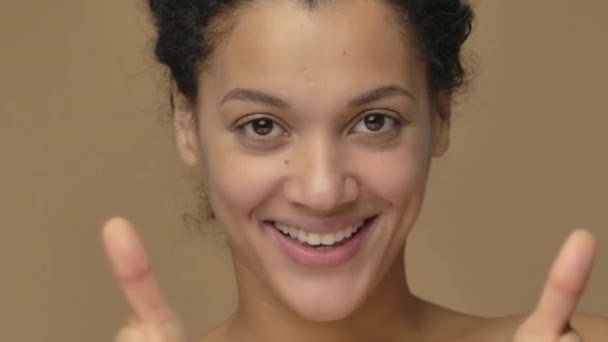 Portrait de beauté de jeune femme afro-américaine souriante et montrant les pouces vers le haut geste. Modèle féminin noir posant sur fond de studio brun. Ferme là. Prêt au ralenti, 4K à 59.94fps. — Video