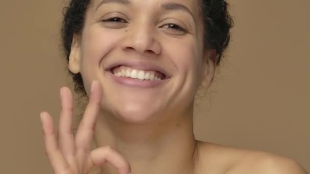 Krásný portrét mladé Afroameričanky, usmívající se, mrkající a ukazující znamení "ok". Black female model pózuje na hnědém pozadí studia. Zavřít. Zpomalení připraveno, 4K při 59,94fps. — Stock video